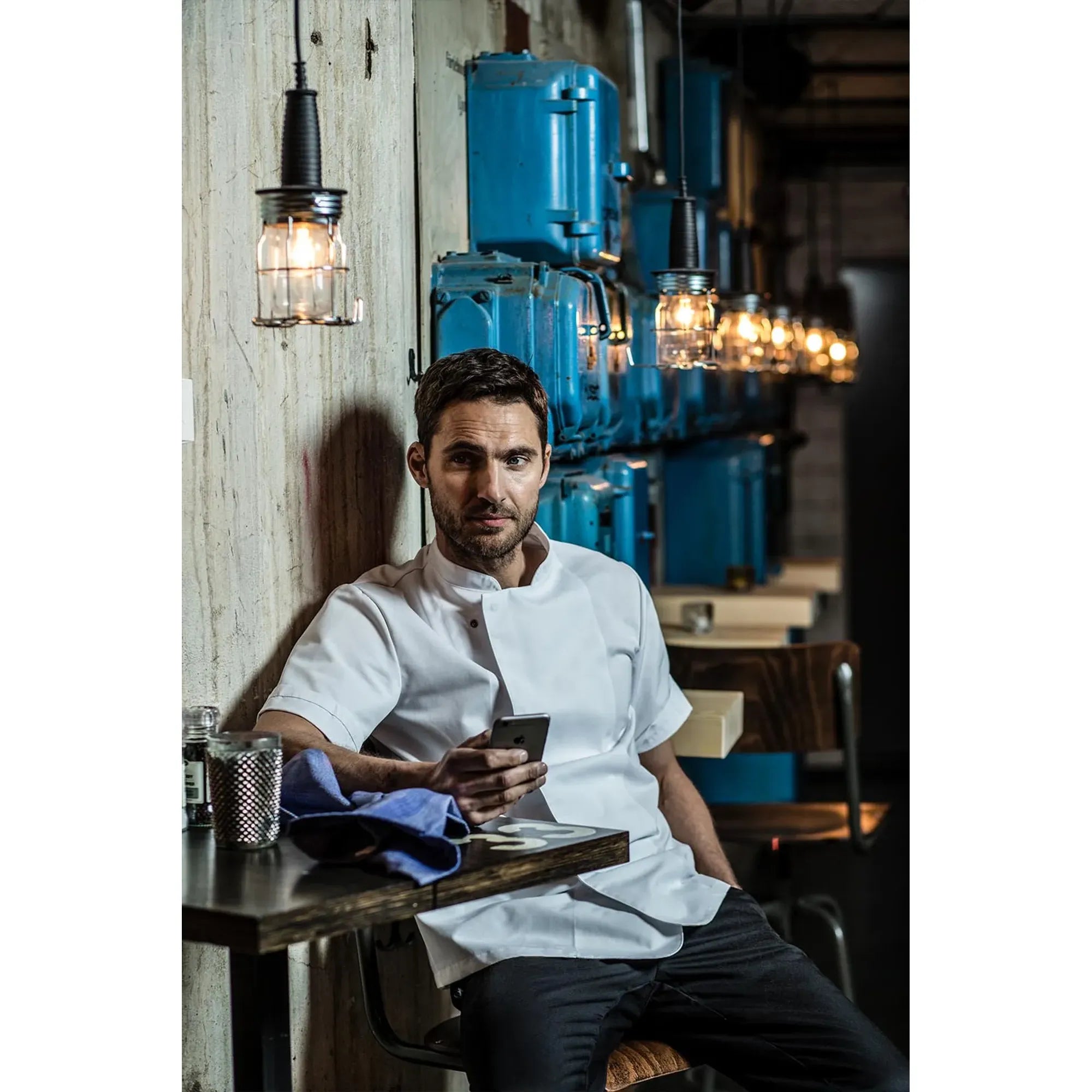 Man in a white double breasted short sleeve chef jacket sitting at a table in a modern interior restaurant