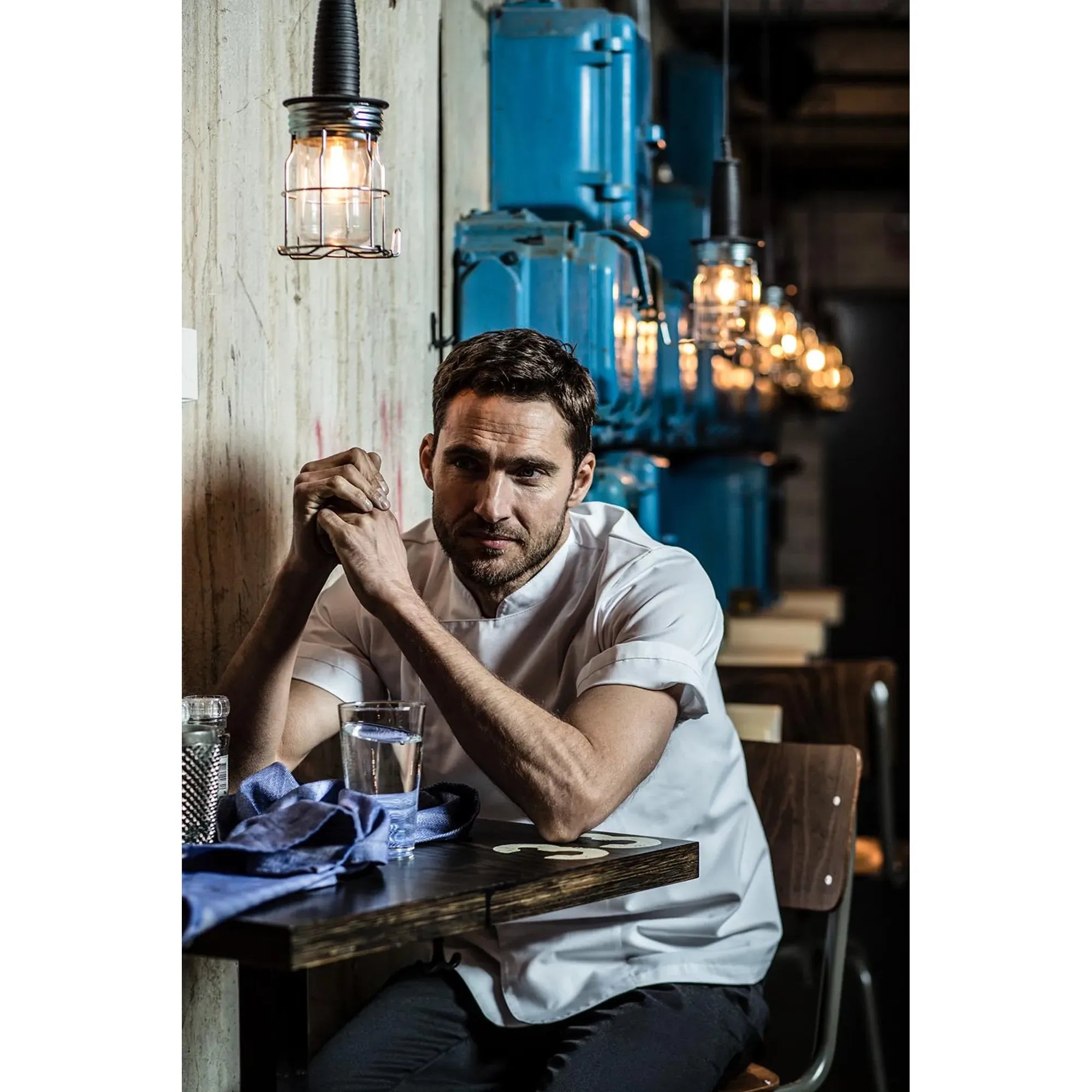 Man sitting at a table in a restaurant with industrial decor, Bring a white chef jacket after a long shift