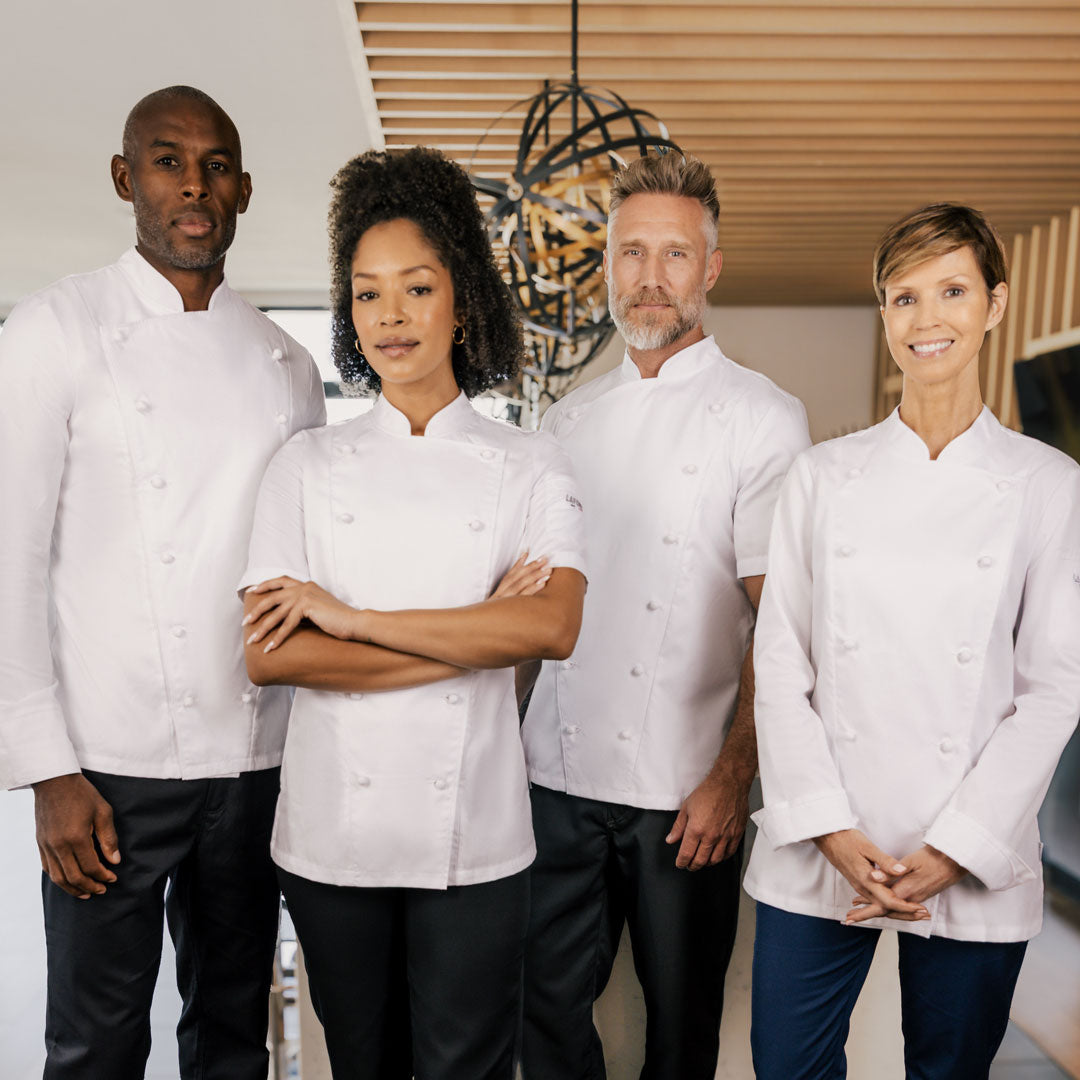 Four executive chefs wearing matching white chef jackets and dark pants are standing together in a modern indoor restaurant before an evening shift.