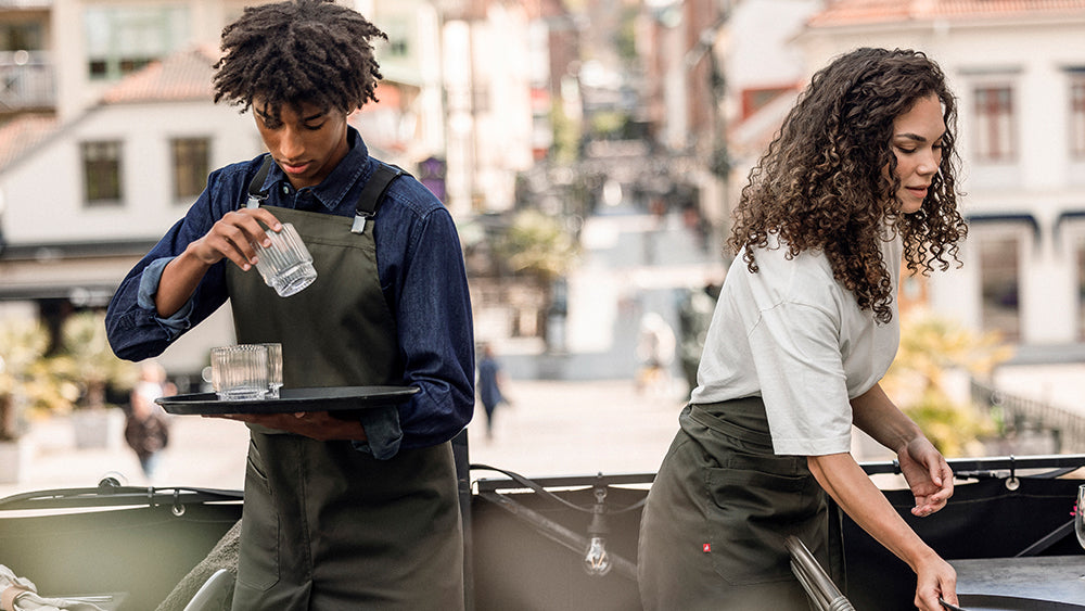 Two restaurant staff members in matching dark green Segers aprons working side by side in a bright, sunlit outdoor cafe setting.