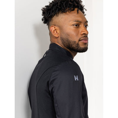 Man wearing a black cooks jacket with an H logo on a light gray background